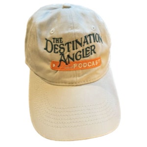 Può includere: Un cappello da baseball beige con il testo "The Destination Angler Podcast" ricamato in verde e arancione.