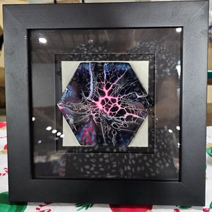Puede incluir: Una pintura abstracta hexagonal enmarcada con colores negros, blancos, azules y rosas que se arremolinan. La pintura está enmarcada en un marco negro con un cristal protector.