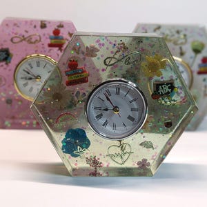 May include: A hexagonal resin clock with a white face and black hands. The clock is decorated with glitter, dried flowers, and small charms, including a heart, a book with the words "Art & Math", and a chalkboard with the letters "ABC".