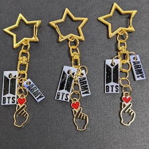 May include: Three gold star-shaped keychains with a chain and charms. Each charm has a black and white glitter background with the text "BTS" and "ARMY" in white. The charms also have a gold finger heart charm.