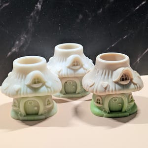 May include: Three small, mushroom-shaped house figurines with a pale, off-white top and a light green base. Each figurine features a rounded roof with heart-shaped details, a small arched window, and a central arched door with a circular handle.