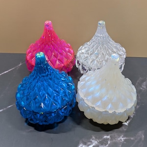 May include: Four decorative lidded jars in vibrant colors: hot pink, clear, blue, and iridescent white. Each jar features a diamond-patterned design and a pointed finial. The jars are displayed on a dark, marbled surface.