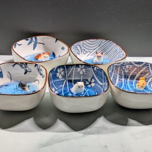 May include: Set of five ceramic bowls with blue and white patterns. Each bowl features a small cat figurine floating in a blue liquid. The bowls have a square shape with rounded corners and a brown rim.