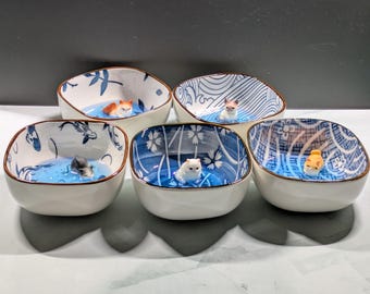 May include: Set of five ceramic bowls with blue and white patterns. Each bowl features a small cat figurine floating in a blue liquid. The bowls have a square shape with rounded corners and a brown rim.