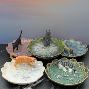 May include: Five scalloped ceramic trinket dishes in various colors, each featuring a small cat figurine and silver leaf flakes. The dishes are pink, green, blue, white, and dark green, with cats in black, gray, tan, orange, and tabby patterns.