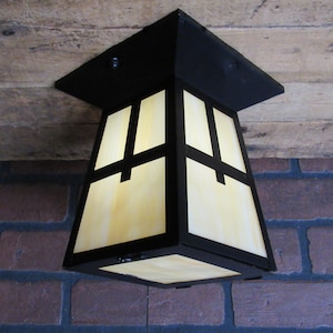 May include: A Craftsman-style ceiling light fixture with a black frame and cream-colored glass panels. The light has a square top and a tapered body, with black cross-shaped accents on the glass. The fixture is mounted on a wooden ceiling.
