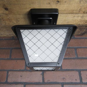 May include: Black outdoor light fixture with a white, diamond-patterned glass panel. The light is mounted on a brick wall and a wooden surface. The fixture has a square base and a tapered, lantern-like shape.