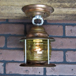May include: A copper and brass ceiling light fixture with a clear glass shade and a yellow bulb. The light is mounted on a wooden beam and has a chain hanging from it.