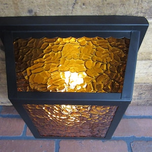 May include: A black metal outdoor wall light fixture with a textured amber glass shade. The light is on and illuminates the glass.