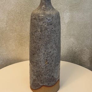 May include: A tall, cylindrical ceramic vase with a narrow neck and a flared rim. The body features a textured, speckled grey and charcoal glaze, while the base is finished in a contrasting smooth, unglazed tan clay with a warm orange-brown tone.