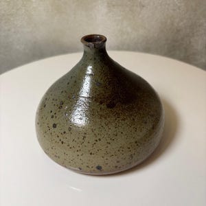 Könnte beinhalten: Kleine, handgefertigte Keramikvase mit rundem Körper und schmalem Hals. Die Vase hat eine gesprenkelte, olivgrüne Glasur, die oben in ein dunkleres Braun übergeht. Die Vase ist etwa 13 cm hoch.