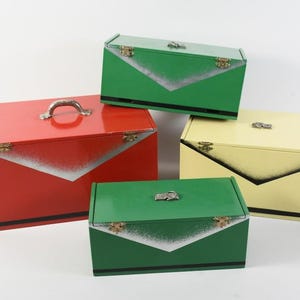 Puede incluir: Un conjunto de cuatro cajas de almacenamiento rectangulares de metal en rojo, amarillo y dos tonos de verde. Cada caja cuenta con tapa abatible, cierres metálicos, un detalle decorativo en forma de V con degradado y una fina franja negra.