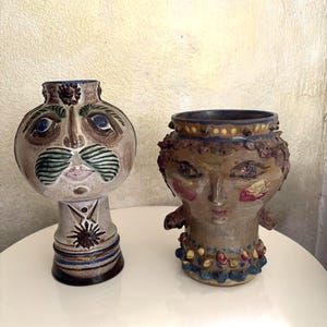 May include: Two ceramic vases shaped like human heads. The left vase is light grey with a prominent green moustache, blue eyes, and a star emblem on the neck. The right vase is brown with textured hair, red cheek accents, and a colourful beaded collar.