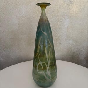 Può includere: Un elegante vaso di vetro con collo sottile e base svasata. Il vaso presenta una sfumatura di colori, che passa da una parte superiore marrone dorato a un corpo verde acqua. Disegni astratti bianchi e fluenti adornano la superficie.