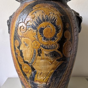 Può includere: Un grande vaso in ceramica caratterizzato da una palette di colori marrone scuro e ocra, con un rilievo raffigurante una figura di profilo che indossa un elmo ornato. Il vaso presenta un bordo largo e svasato e una superficie ruvida e incisa.
