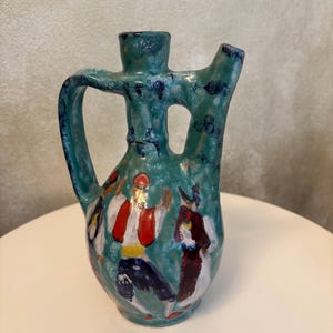 May include: A turquoise ceramic pitcher with a mottled glaze finish, featuring a handle and a spout. The body of the pitcher is decorated with hand-painted figures in red, white, yellow, and dark blue attire, depicted in a dynamic dancing pose.