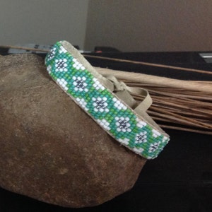 May include: A green and white beaded bracelet with a floral pattern. The bracelet is lying on a brown rock.
