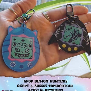 May include: Two acrylic keyrings featuring retro-style digital pet designs. One is blue with a cat-like shape, the other is black with a bat-like shape. Both have digital screens displaying pixelated graphics. The text "KPOP DEMON HUNTERS" and "DERPY & SUSSIE TAMAGOTCHI ACRYLIC KEYRINGS" is visible.