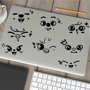 May include: A silver laptop with black cartoon faces with different expressions on the lid. The faces include a deer, a pig, a bird, a cat, and a few other faces with different expressions.
