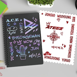 May include: A black notebook with a spiral binding featuring colorful stickers of the K-pop group A.C.E. The stickers include the group's name, member names, and various symbols. A white sheet with red stickers of the K-pop group ATEEZ is next to the notebook. The stickers include the group's name, member names, and various symbols.