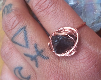 May include: A copper wire wrapped ring with a black obsidian stone. The ring is on a finger with tattoos.