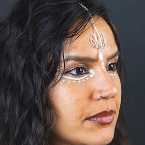 Pode incluir: Uma mulher com cabelo comprido e preto usando uma tiara de flor de lótus prateada e um pequeno anel de nariz roxo. Ela tem pintura facial branca e dourada com pontos brancos.