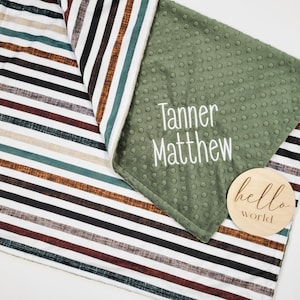May include: A green and white striped baby blanket with a green minky side. The blanket has the names "Tanner Matthew" embroidered on the green side. A wooden sign with the text "hello world" is on the blanket.