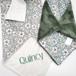 Peut inclure: Une couverture pour b&eacute;b&eacute; &agrave; motifs floraux verts et blancs avec un c&ocirc;t&eacute; doux et pelucheux et un c&ocirc;t&eacute; blanc avec le nom "Quincy" brod&eacute; en vert.