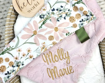May include: A pink and white floral baby blanket with a pink minky backing. The blanket is personalized with the name "Molly Marie" embroidered in gold thread. A wooden sign with the text "hello world" is also in the image.
