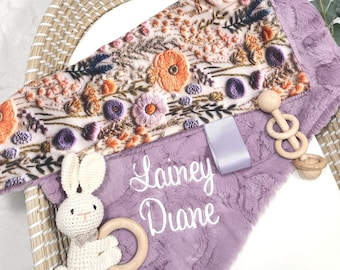 May include: A purple plush baby blanket with a floral embroidered design and the name "Lainey Diane" embroidered on it. The blanket is paired with a crocheted bunny rattle and a wooden teether.