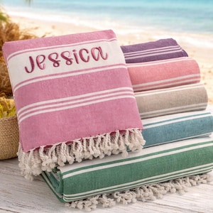 Puede incluir: Una pila de toallas de playa a rayas de varios colores, incluyendo rosa, morado y verde. La toalla superior es rosa y est&aacute; bordada con el nombre "Jessica". Cada toalla tiene rayas blancas y un borde con flecos. El fondo muestra una playa.