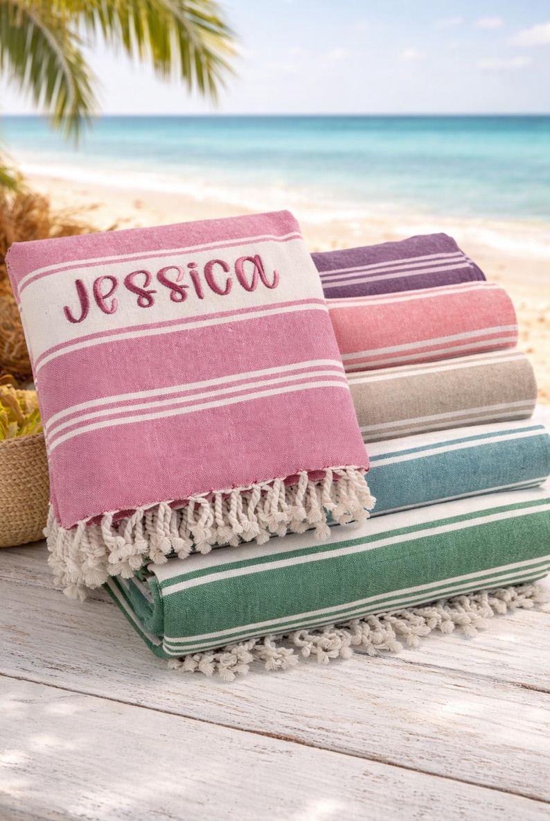 Puede incluir: Una pila de toallas de playa a rayas de varios colores, incluyendo rosa, morado y verde. La toalla superior es rosa y est&aacute; bordada con el nombre "Jessica". Cada toalla tiene rayas blancas y un borde con flecos. El fondo muestra una playa.