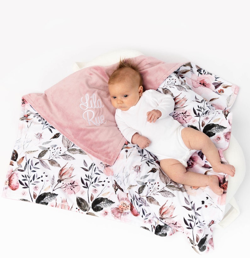 May include: A pink and white baby blanket with a floral pattern. The blanket has a soft, plush side and a cotton side. The blanket is personalized with the name "Lila Rue" embroidered on the pink side.