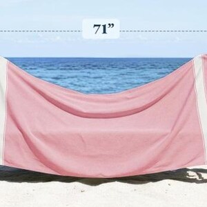 Puede incluir: Una toalla de playa rosa con rayas blancas y flecos, sobre una silla de playa de madera. La toalla mide 180 cm de largo y 99 cm de ancho. El fondo muestra el mar y el cielo.