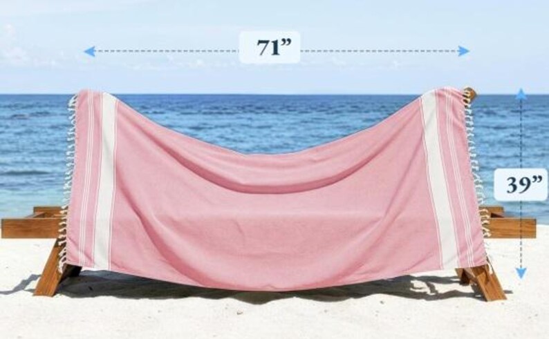 Puede incluir: Una toalla de playa rosa con rayas blancas y flecos, sobre una silla de playa de madera. La toalla mide 180 cm de largo y 99 cm de ancho. El fondo muestra el mar y el cielo.