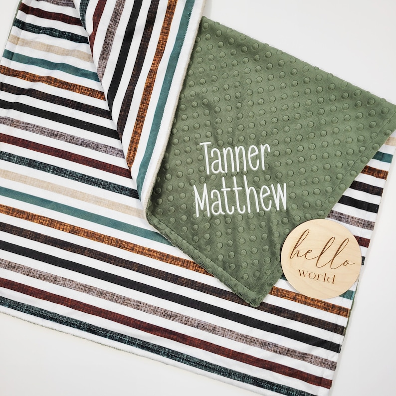 May include: A green and white striped baby blanket with a green minky side. The blanket has the names "Tanner Matthew" embroidered on the green side. A wooden sign with the text "hello world" is on the blanket.