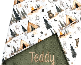 Puede incluir: Una manta para bebé con temática de camping. La capa superior presenta un fondo blanco con ilustraciones de tiendas de campaña, árboles, fogatas, osos y huellas de patas. La capa inferior es verde oliva con un patrón texturizado de puntos. El nombre "Teddy" está bordado en beige.