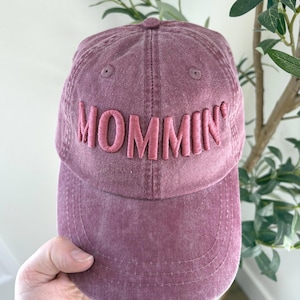 May include: A dusty rose baseball cap with the word "MOMMIN'" embroidered in a lighter pink. The cap is held by a hand and has a curved brim. The background includes greenery.