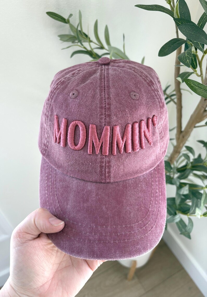 May include: A dusty rose baseball cap with the word "MOMMIN'" embroidered in a lighter pink. The cap is held by a hand and has a curved brim. The background includes greenery.