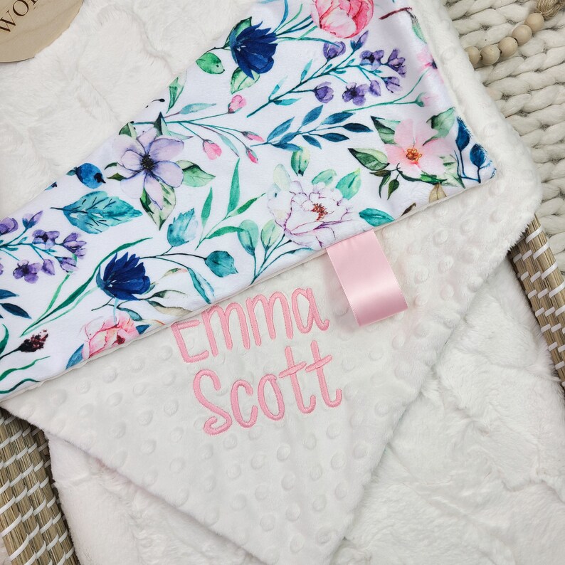 May include: White baby blanket with a floral print on one side and the name "Emma Scott" embroidered on the other side.