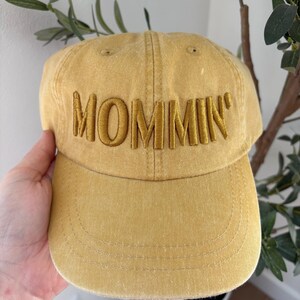 May include: Mustard yellow baseball cap with the word "MOMMIN'" embroidered across the front in a matching color. The cap has a curved brim and a classic six-panel design, suitable for casual wear.