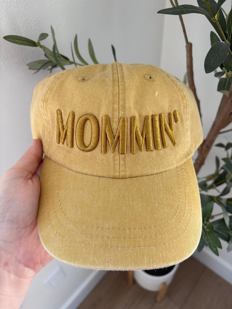 May include: Mustard yellow baseball cap with the word "MOMMIN'" embroidered across the front in a matching color. The cap has a curved brim and a classic six-panel design, suitable for casual wear.