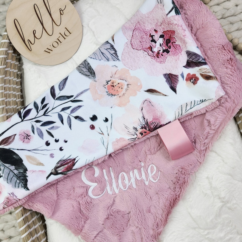 May include: A pink and white baby blanket with a floral print and the name "Ellonie" embroidered on it. The blanket is folded in half and has a small pink tag attached to it.
