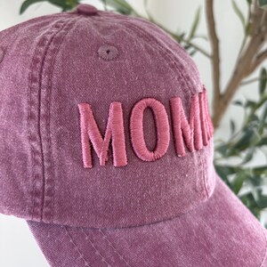 May include: A dusty rose baseball cap with the word "MOM" embroidered in a matching pink thread. The cap has a curved brim and a fabric-covered button on top. The hat is made of a soft, textured material.