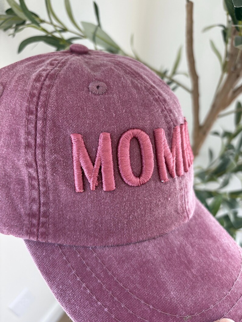 May include: A dusty rose baseball cap with the word "MOM" embroidered in a matching pink thread. The cap has a curved brim and a fabric-covered button on top. The hat is made of a soft, textured material.
