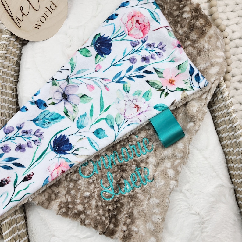 May include: A white and brown baby blanket with a floral print and the name "Emmalie Lisette" embroidered in teal on the brown side.