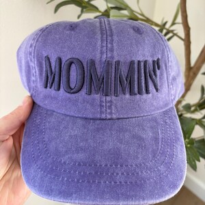 May include: A purple baseball cap with the word "MOMMIN" embroidered in navy blue. The cap has a curved brim and a slightly faded appearance, suggesting a vintage or worn look. The cap is made of a soft fabric.
