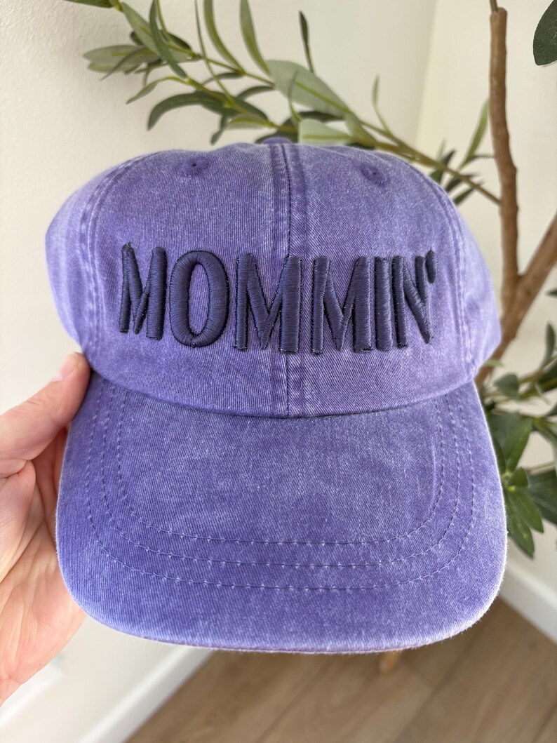 May include: A purple baseball cap with the word "MOMMIN" embroidered in navy blue. The cap has a curved brim and a slightly faded appearance, suggesting a vintage or worn look. The cap is made of a soft fabric.