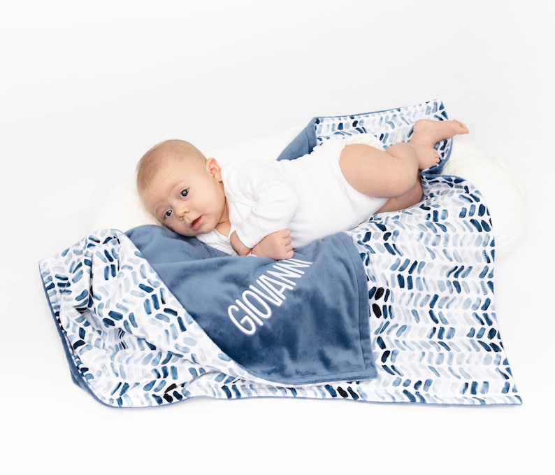 May include: A blue and white baby blanket with a chevron pattern on one side and a solid blue side with the name "Giovanni" embroidered in white. A baby is lying on the blanket.