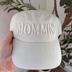 May include: A light gray baseball cap with the word "MOMMIN'" embroidered in a lighter shade of gray across the front. The cap has a curved brim and a classic six-panel design. The background includes greenery.